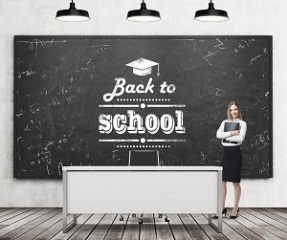 A beautiful girl with black document folder and teacher's desk in a modern school. A black chalkboard on the wall with written down phrase - Back to school. Three black ceiling lights.